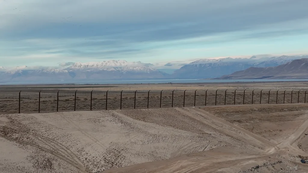 The 45-foot perimeter wind fence at Bayview Landfill, its tilted upper arms angled inward over the site, with Utah Lake and the snow-capped Wasatch Range beyond.